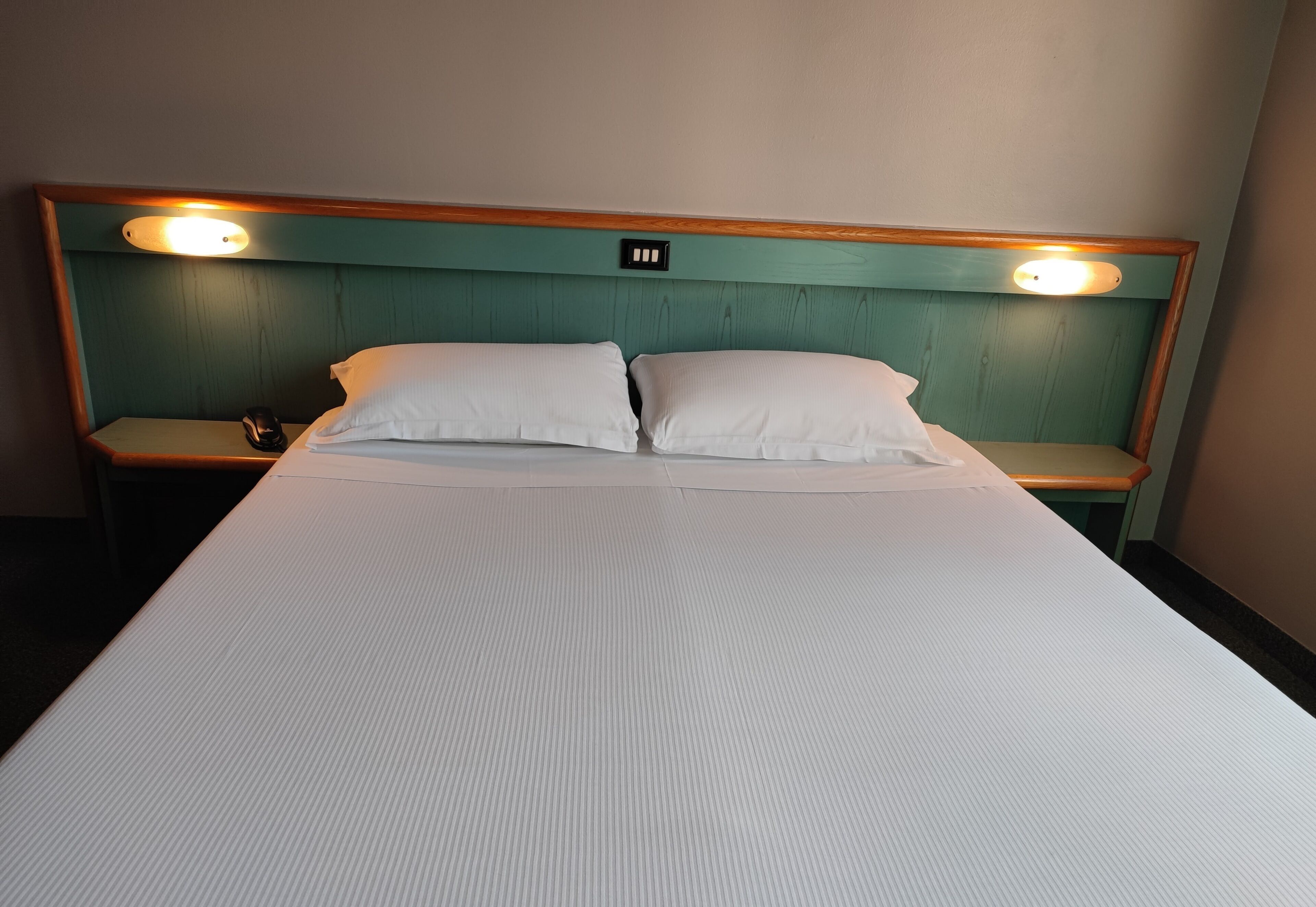 A hotel room with a single bed, two pillows, a wooden headboard with built-in lighting, and a bedside table with a phone and a remote control.