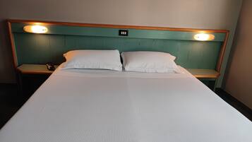 A hotel room with a single bed, two pillows, a wooden headboard with built-in lighting, and a bedside table with a phone and a remote control.