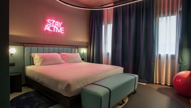 A modern bedroom with a bed, bench, neon sign, and curtains. - Stay Hotel (Brunello)