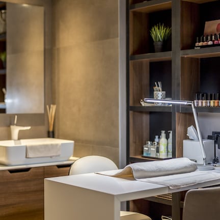 A modern bathroom with a white sink, a mirror, a vanity with a towel, a desk with a lamp, and shelves with toiletries.