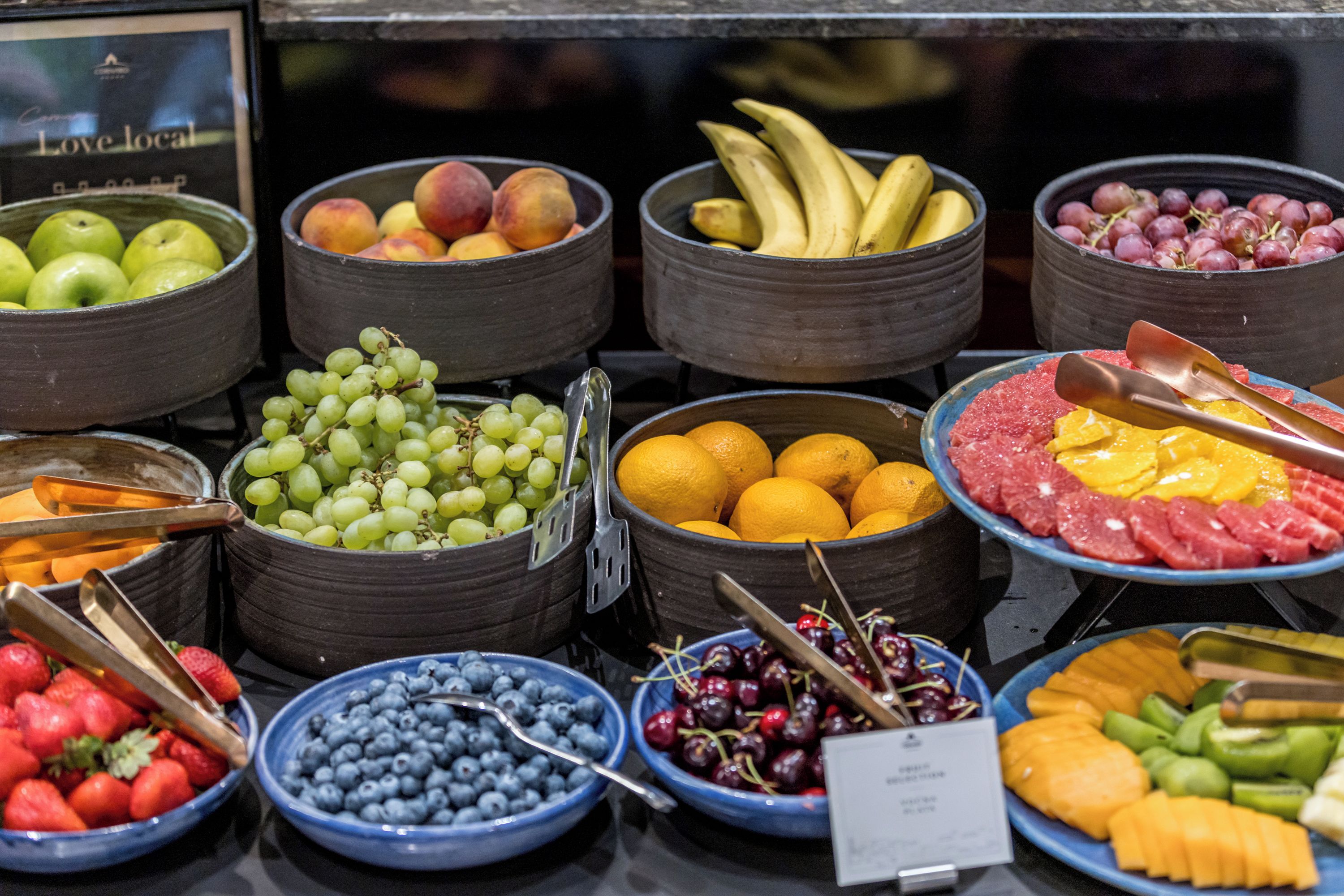 Diversas frutas e legumes dispostos em tigelas e cestas, algumas com pinças para servir.