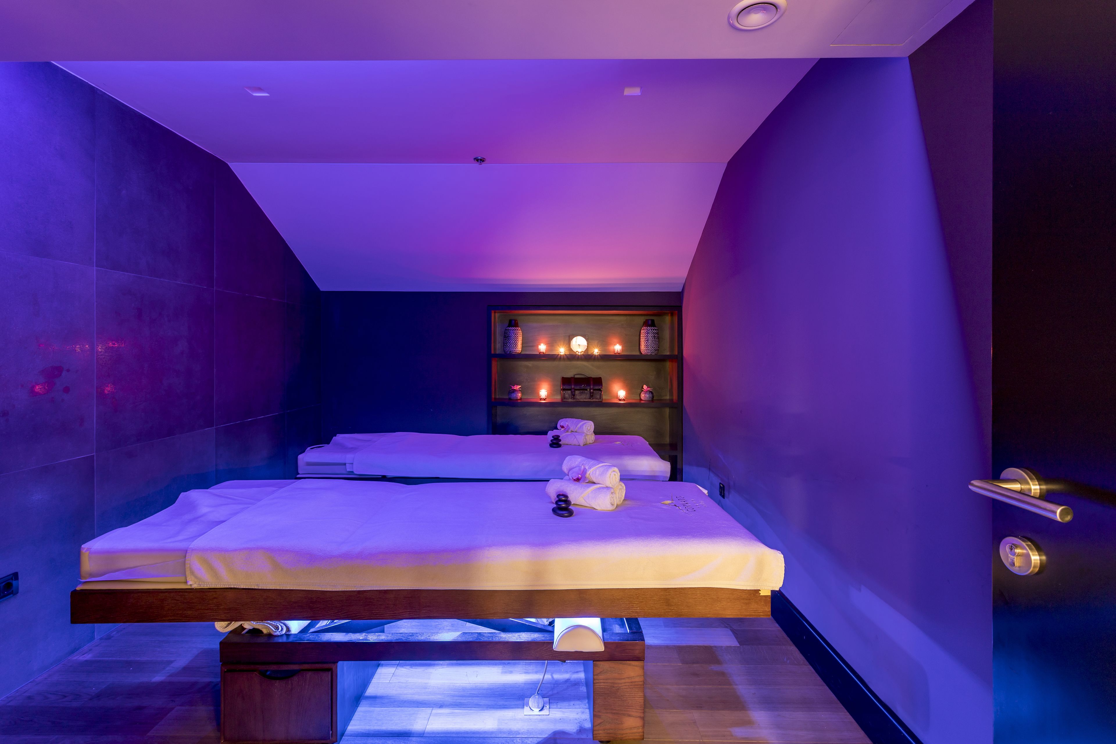 A modern spa room with two beds, a massage table, and a shelf with candles.