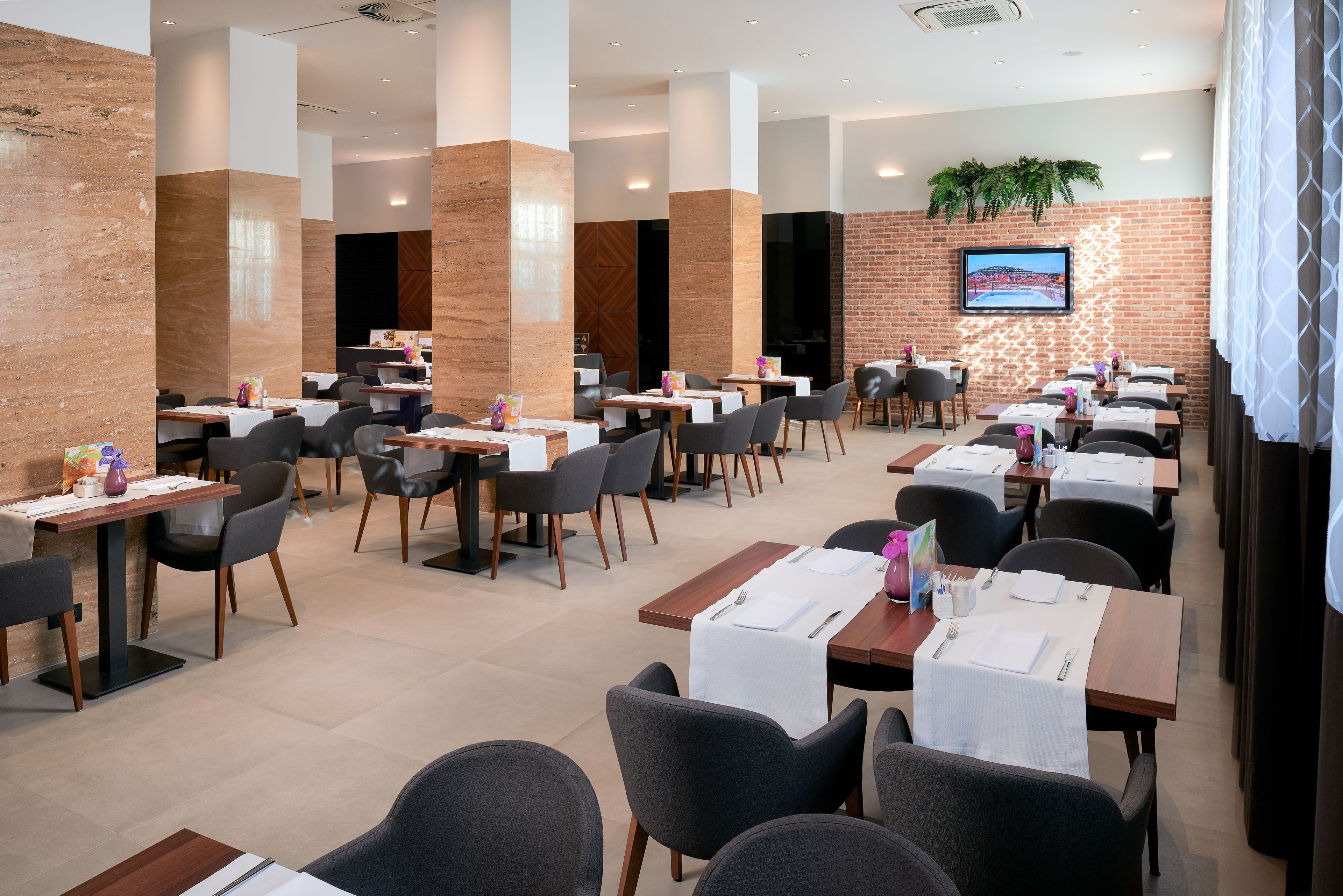 A modern dining area with multiple tables and chairs, a brick wall, and a mounted TV screen.