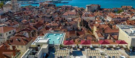 Uma área de piscina no terraço, com várias espreguiçadeiras e mesas, rodeada por edifícios com telhados de cerâmica vermelha.