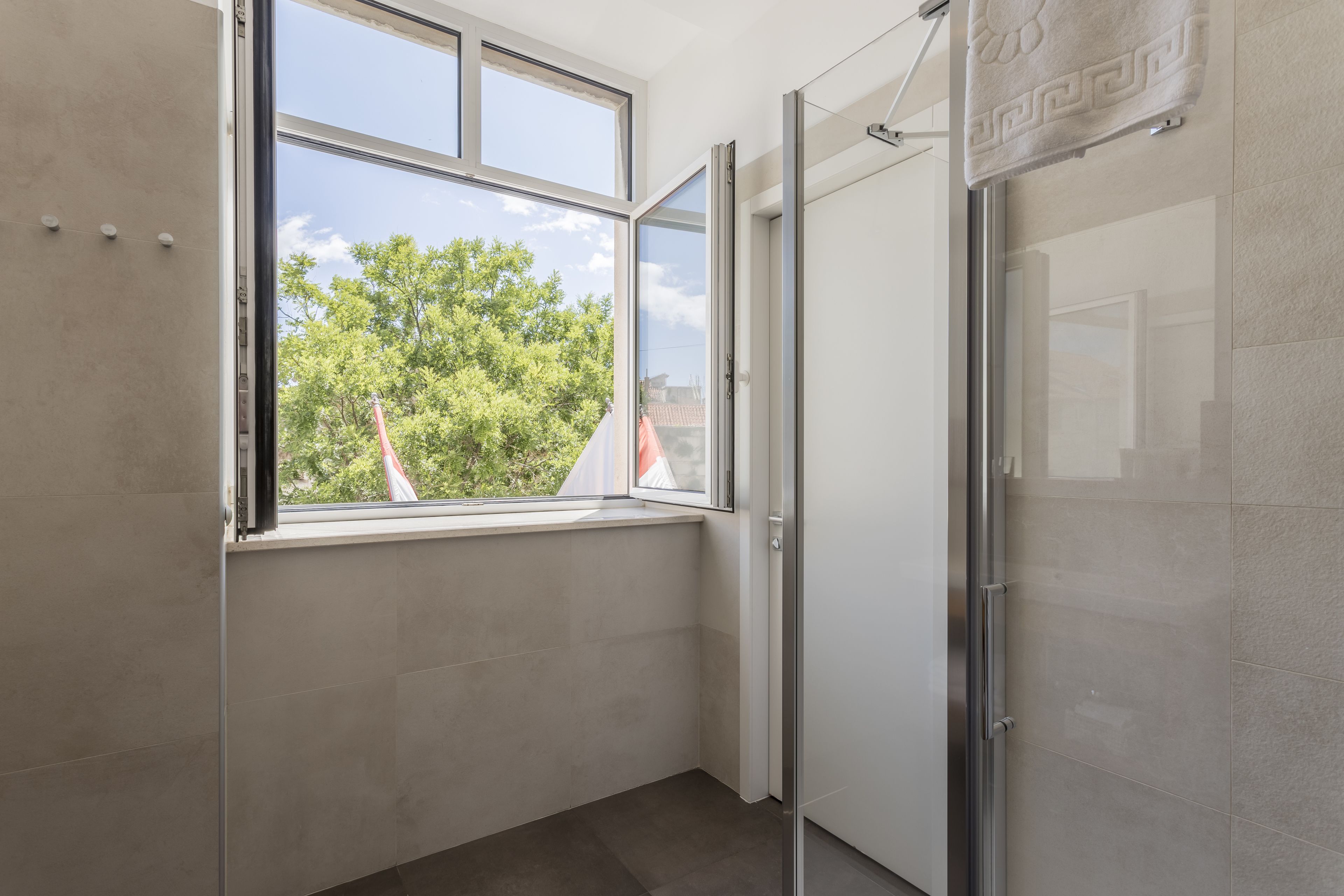 A modern bathroom with a large window, a glass shower enclosure, and a view of trees and buildings outside.