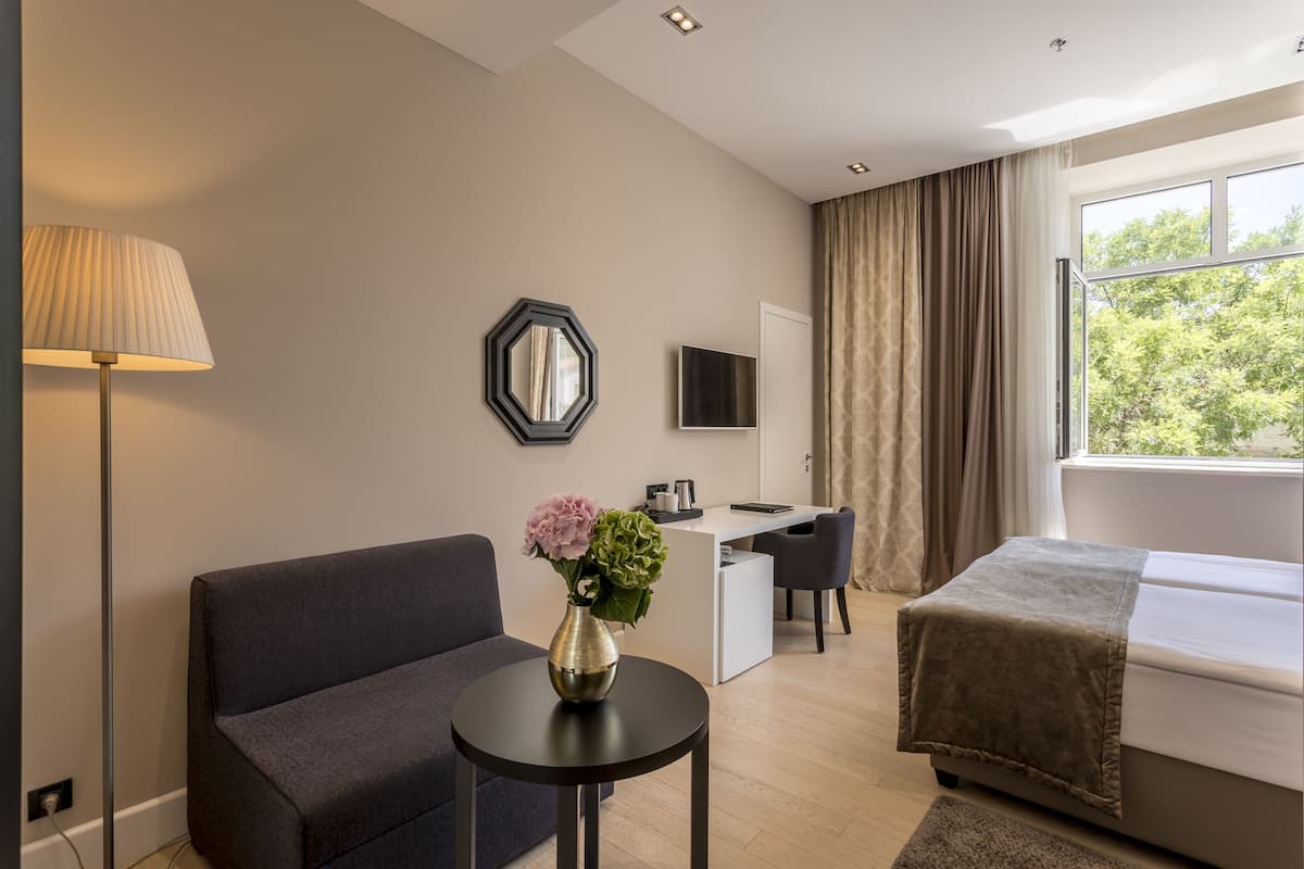 A modern hotel room with a sofa, a small table with a vase of flowers, a desk, a bed, and a large window with curtains.