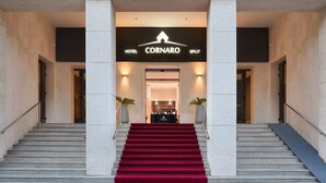 En hotellentrén med en röd matta som löper uppför en trappa, en receptionsdisk och en skylt med texten ”Hotel Cornaro Split”.