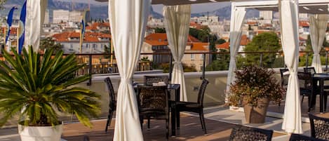Une terrasse extérieure dotée de parasols blancs, de meubles en osier, de plantes en pot et offrant une vue sur un paysage urbain avec des montagnes en arrière-plan.