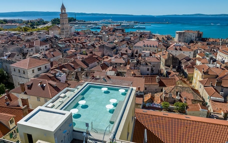 A rooftop pool with multiple sun loungers, surrounded by historic buildings with red-tiled roofs and a prominent tower in the background.
