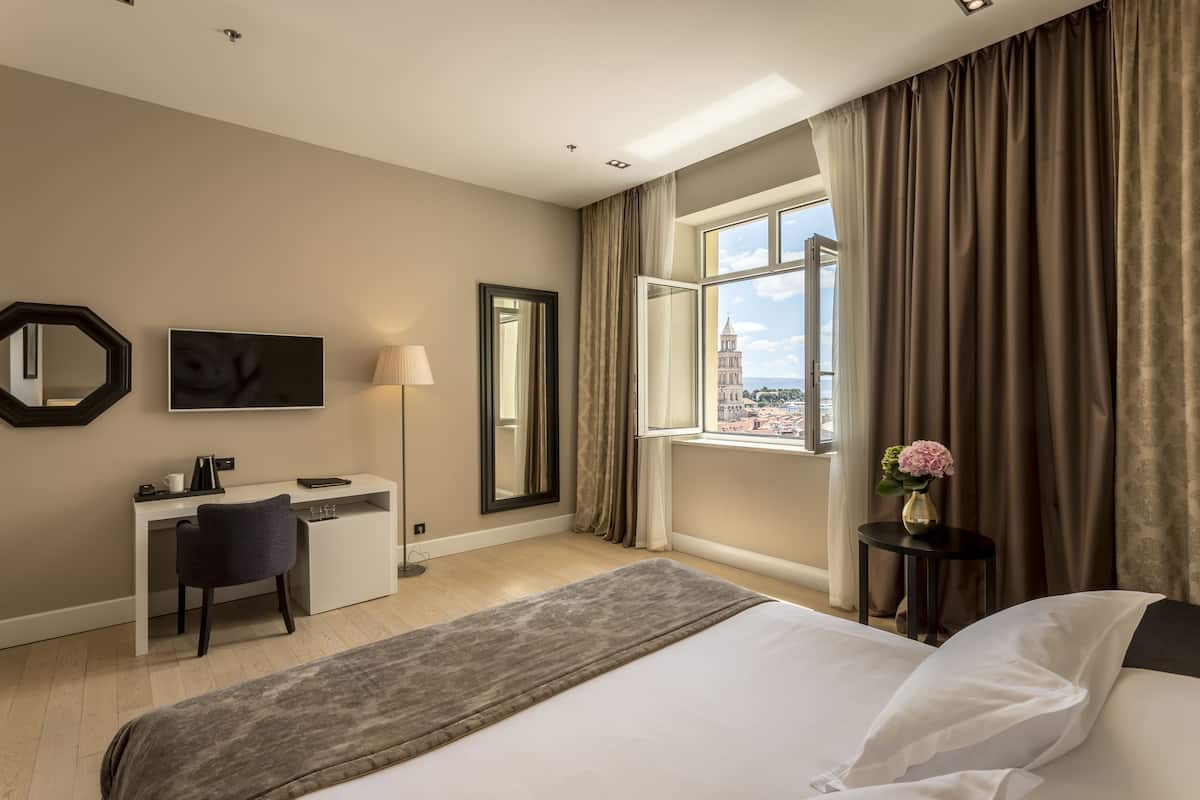 A hotel room with a bed, a desk, a chair, a TV, a window with curtains, and a vase with flowers.