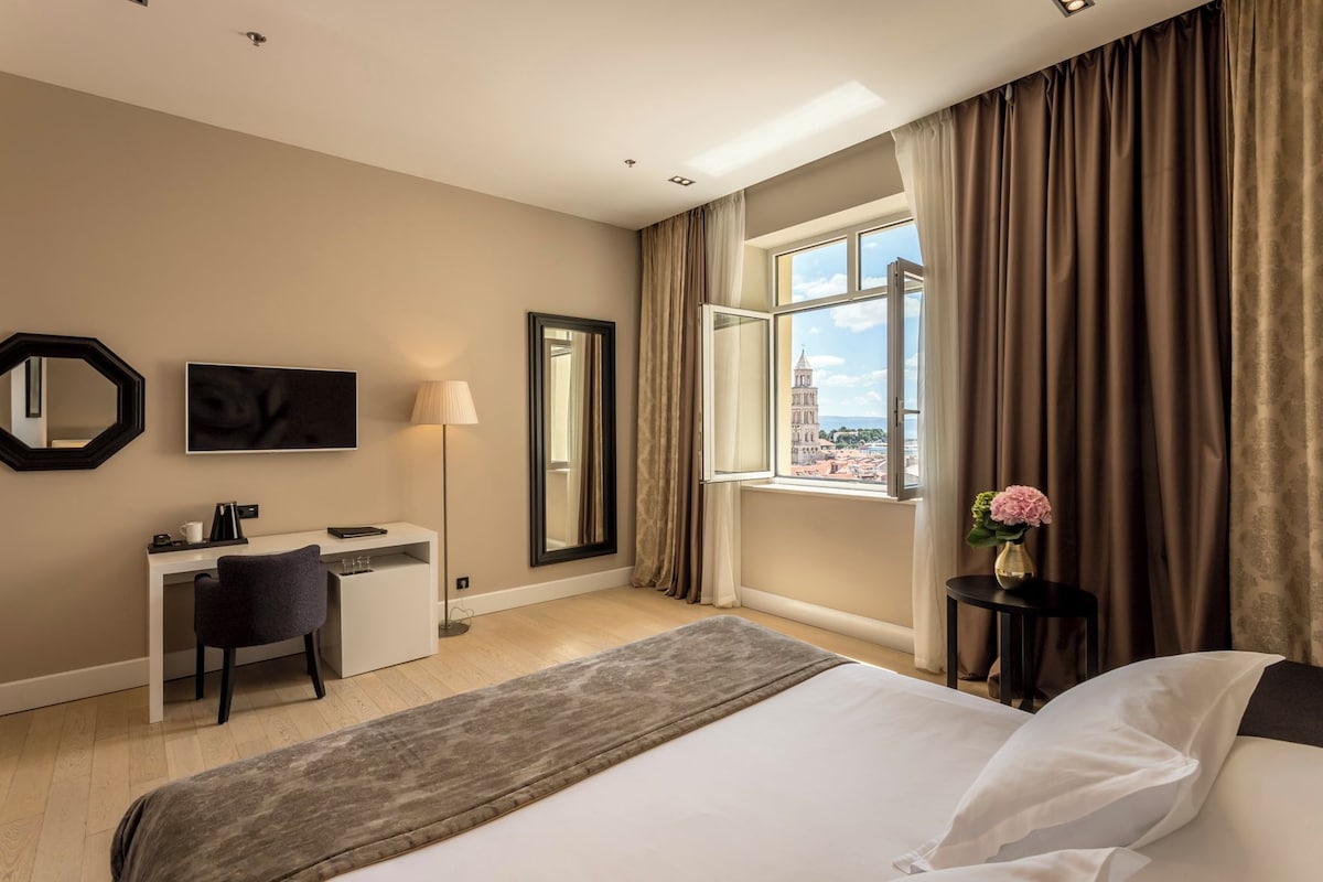 Habitación de hotel con cama, escritorio, silla y un ventanal con cortinas.