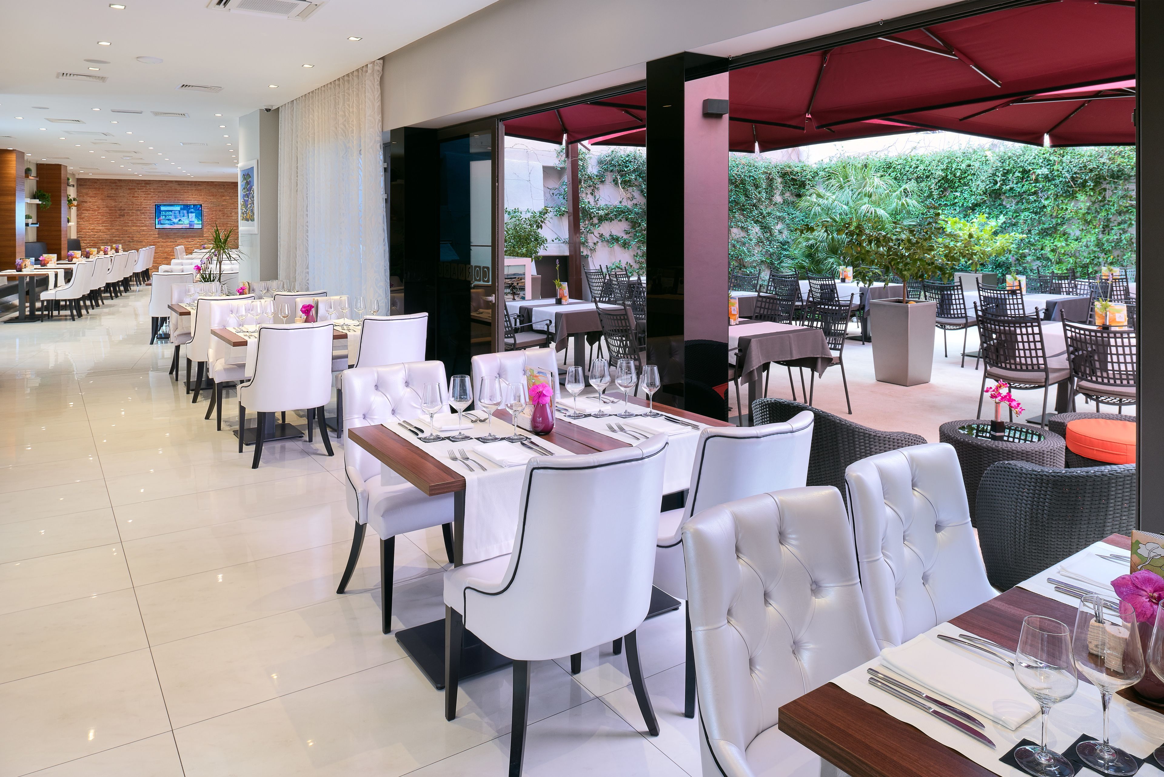 Um restaurante com cadeiras e mesas brancas, uma tela de TV e vista para o verde lá fora.