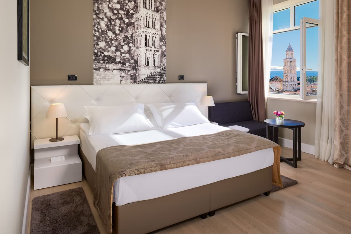 A hotel room with a large bed, a nightstand, a small table with a lamp, a view of a tower through a window, and a painting on the wall.