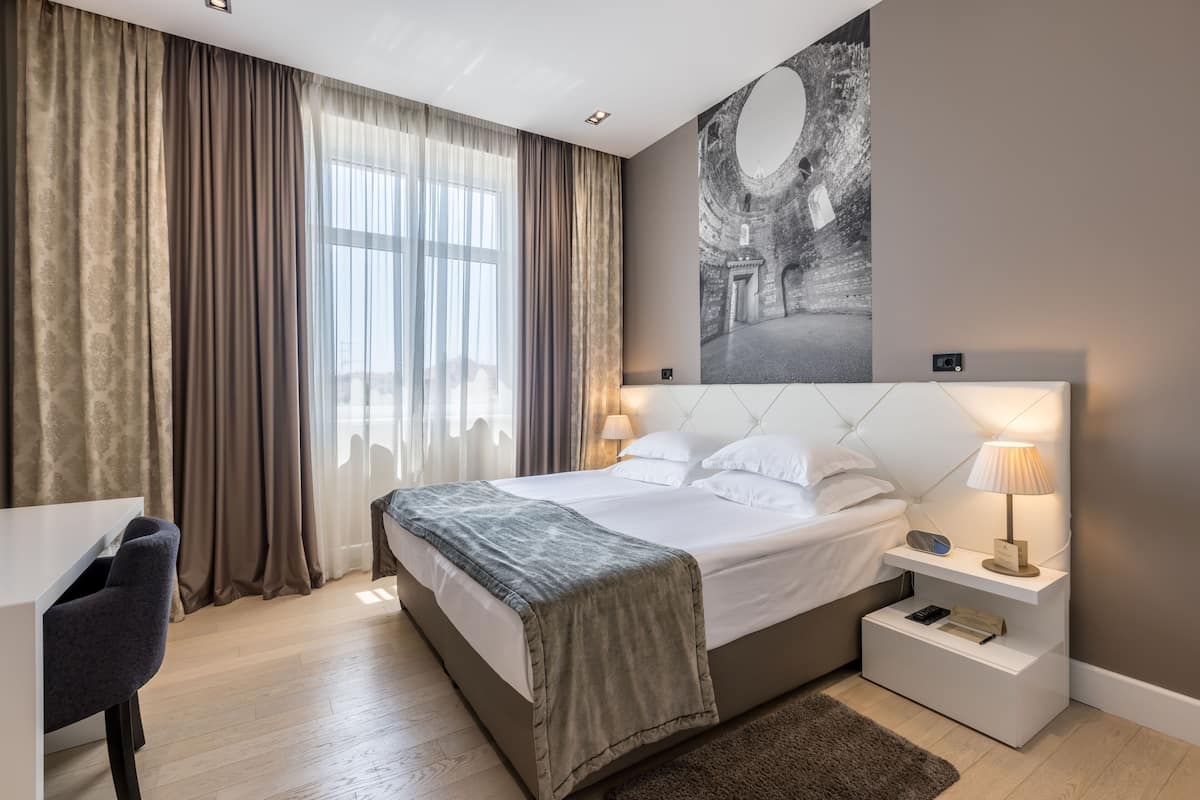 A modern hotel room with a large bed, bedside tables, a desk, and a view of a cityscape through the window.