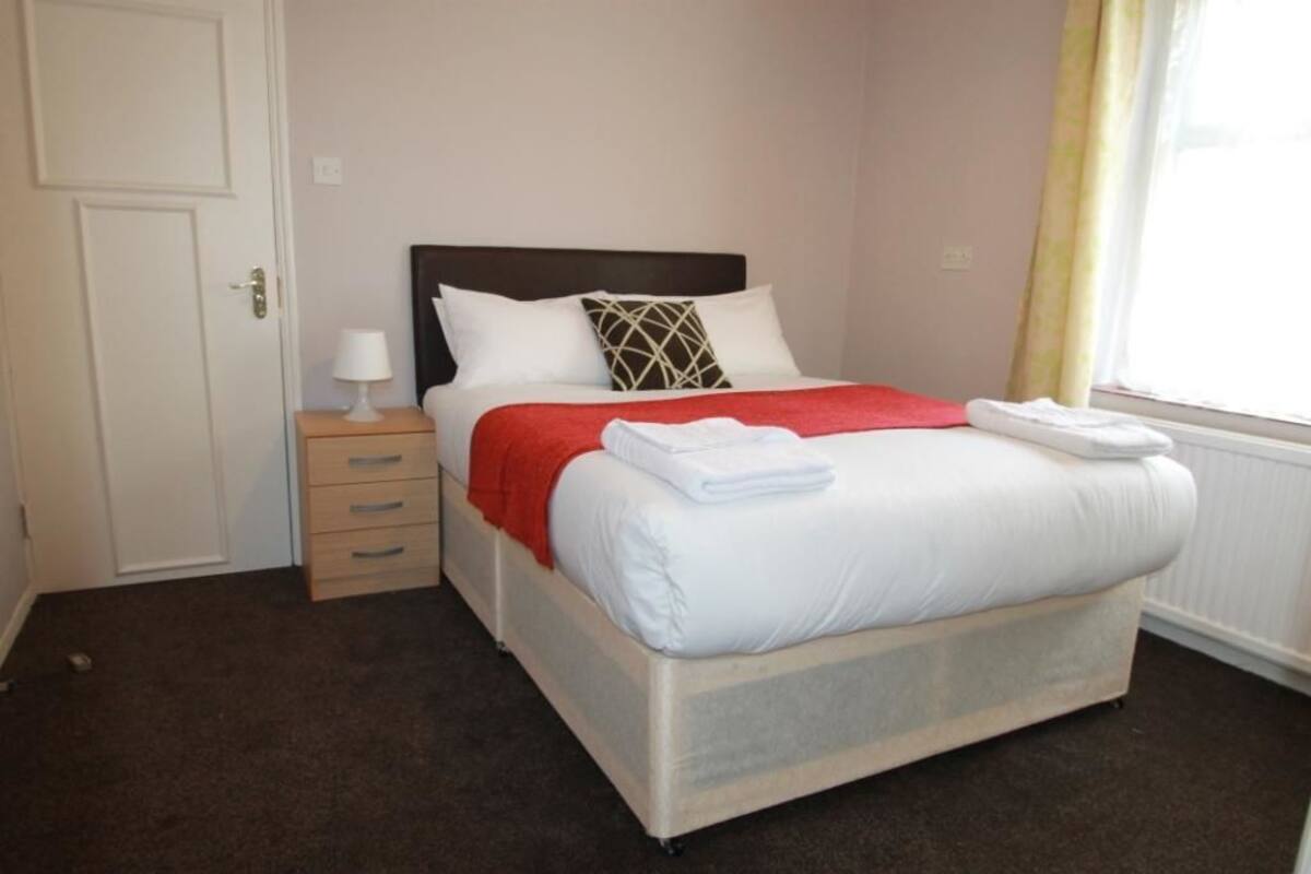 A neatly made bed with white linens and a red blanket, a bedside table with a lamp, and a window with curtains.