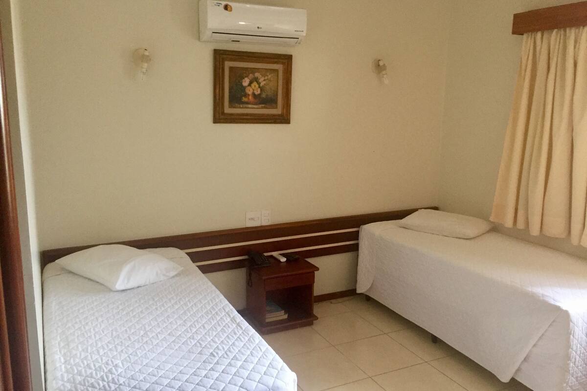 A hotel room with two single beds, a small wooden nightstand, a wall-mounted air conditioner, and a framed picture above the beds.