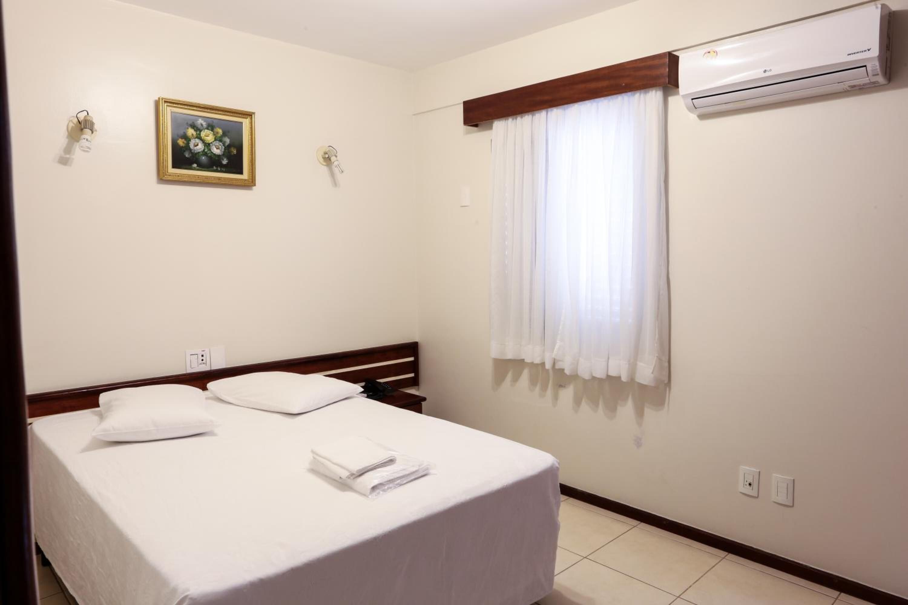 A hotel room with a single bed, a nightstand, an air conditioning unit, a window with curtains, and a framed picture on the wall.