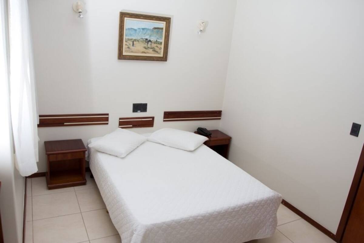 A single bed with a white bedspread, two bedside tables, a nightstand, a painting on the wall, and a telephone on the nightstand.