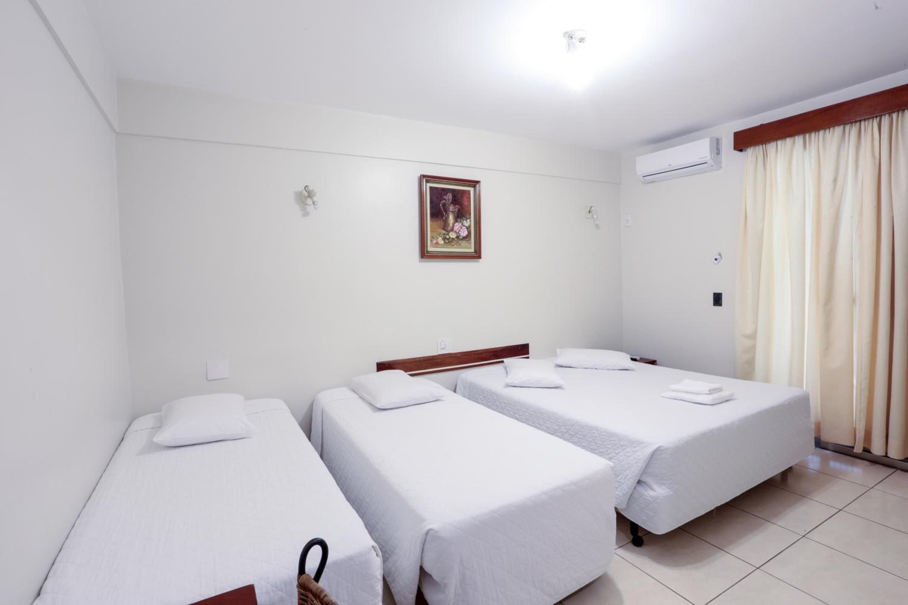 Habitación de hotel con tres camas individuales, un cabecero de madera, un aire acondicionado de pared y un cuadro enmarcado sobre las camas.