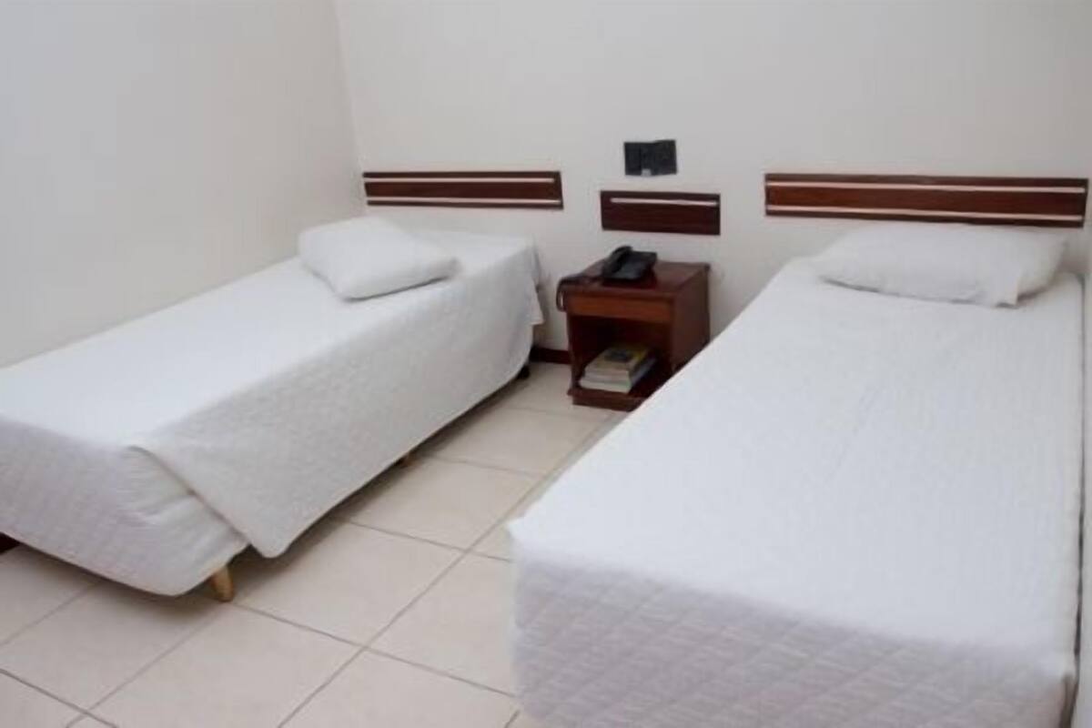 A hotel room with two single beds, a nightstand with a phone and books, and a small wall-mounted shelf.