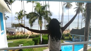 A person standing on a balcony with a view of a pool and palm trees. - Costarena Beach Hotel (Las Terrenas)