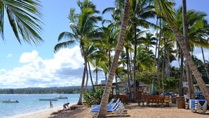 A beach with palm trees, a beachside restaurant, and lounge chairs. - Costarena Beach Hotel (Las Terrenas)