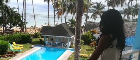 A balcony with a view of a swimming pool, palm trees, and a beach.