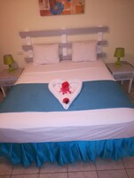 Una cama bien hecha con una toalla en forma de corazón como detalle central, una colcha azul y cortinas a juego.