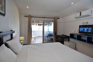 Habitación de hotel con cama, escritorio con televisor, refrigerador pequeño y puerta corrediza de vidrio que da a un espacio exterior con sillas.