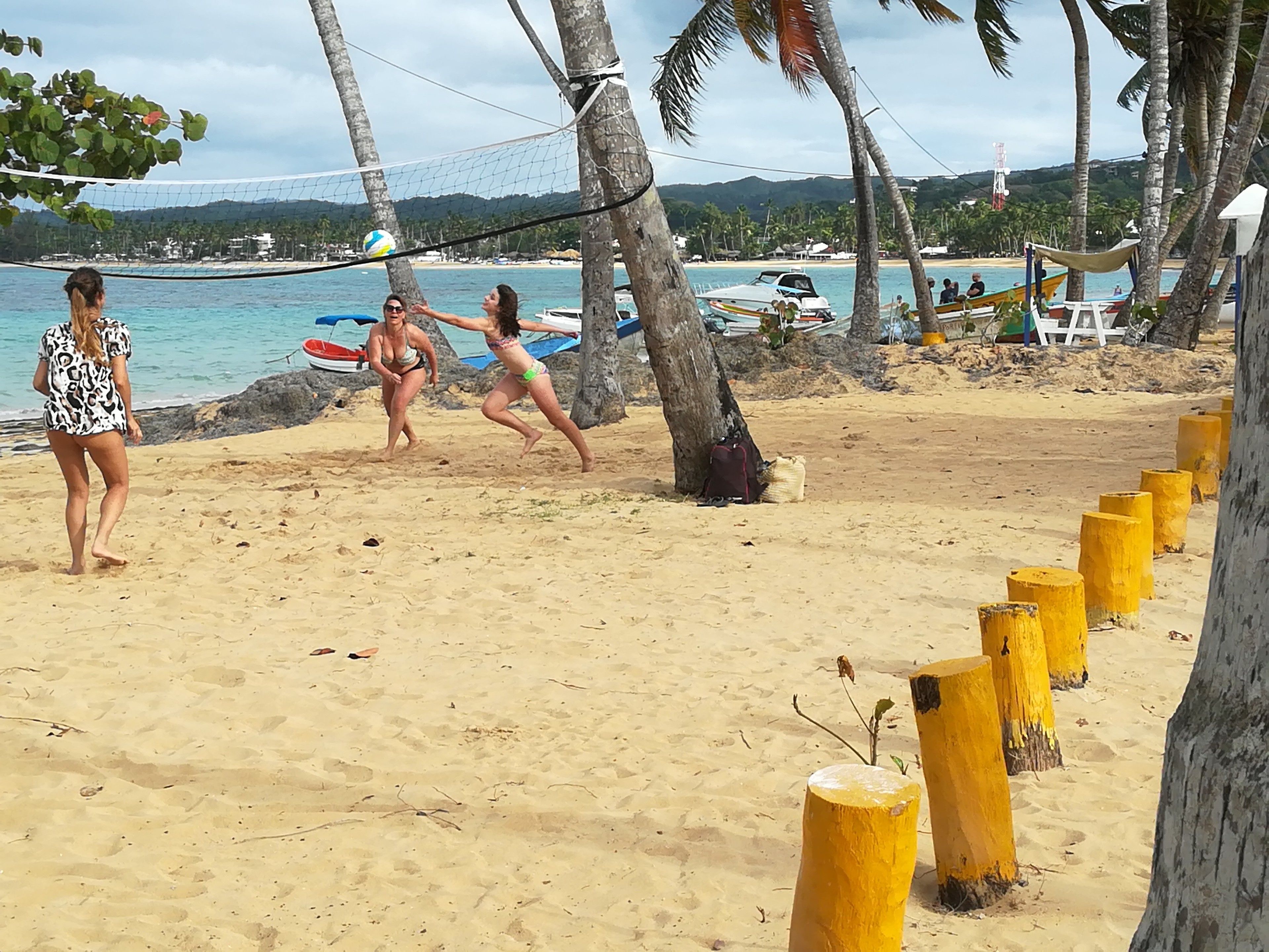 Partido de voleibol de playa con palmeras y barcos al fondo.