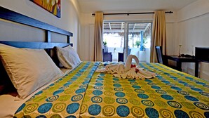 A hotel room with a bed, a headboard, a painting, a window with curtains, and a view of the outside. - Costarena Beach Hotel (Las Terrenas)