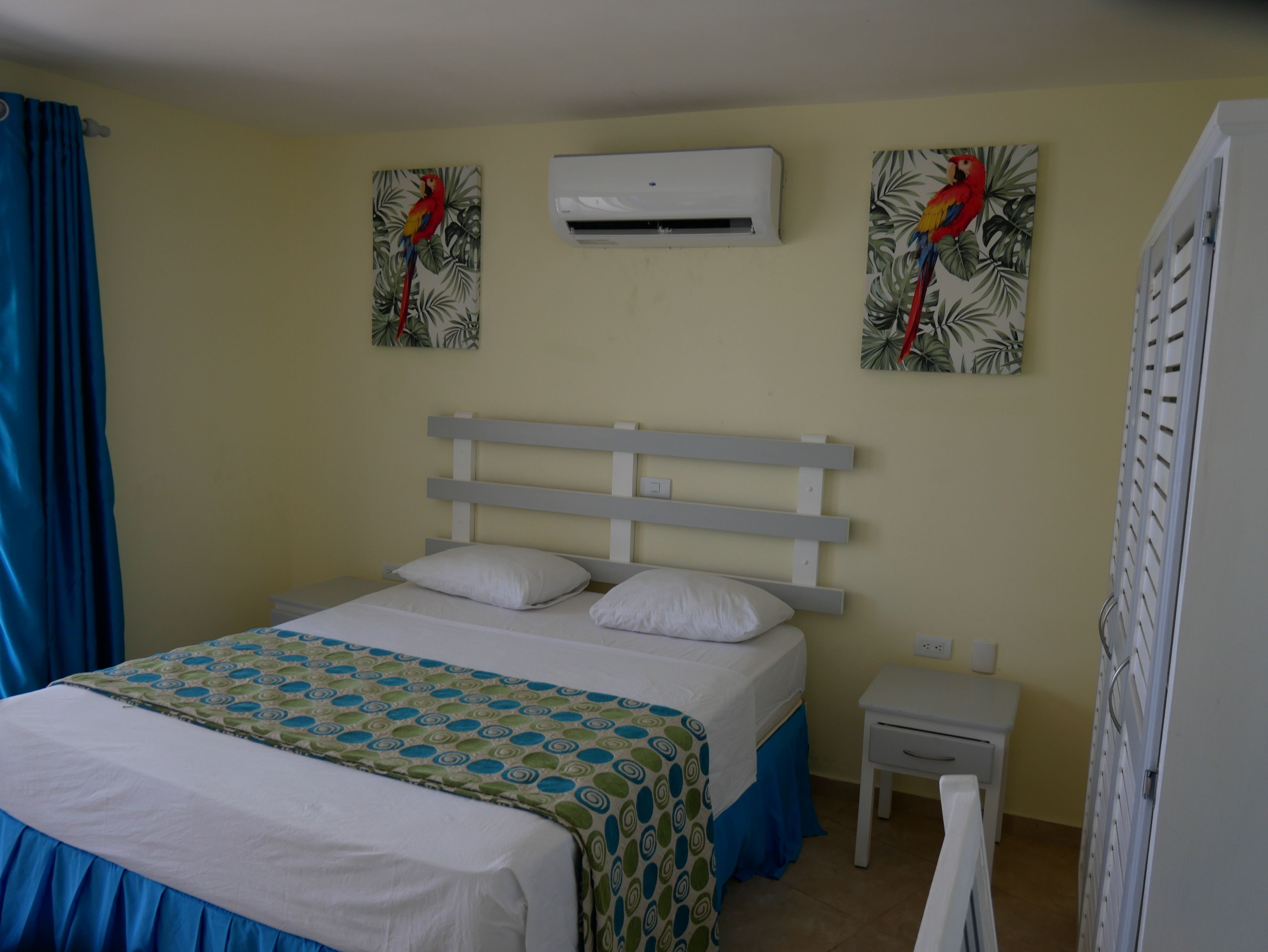 A bedroom with a bed, a nightstand, an air conditioner, and two wall-mounted paintings of parrots.