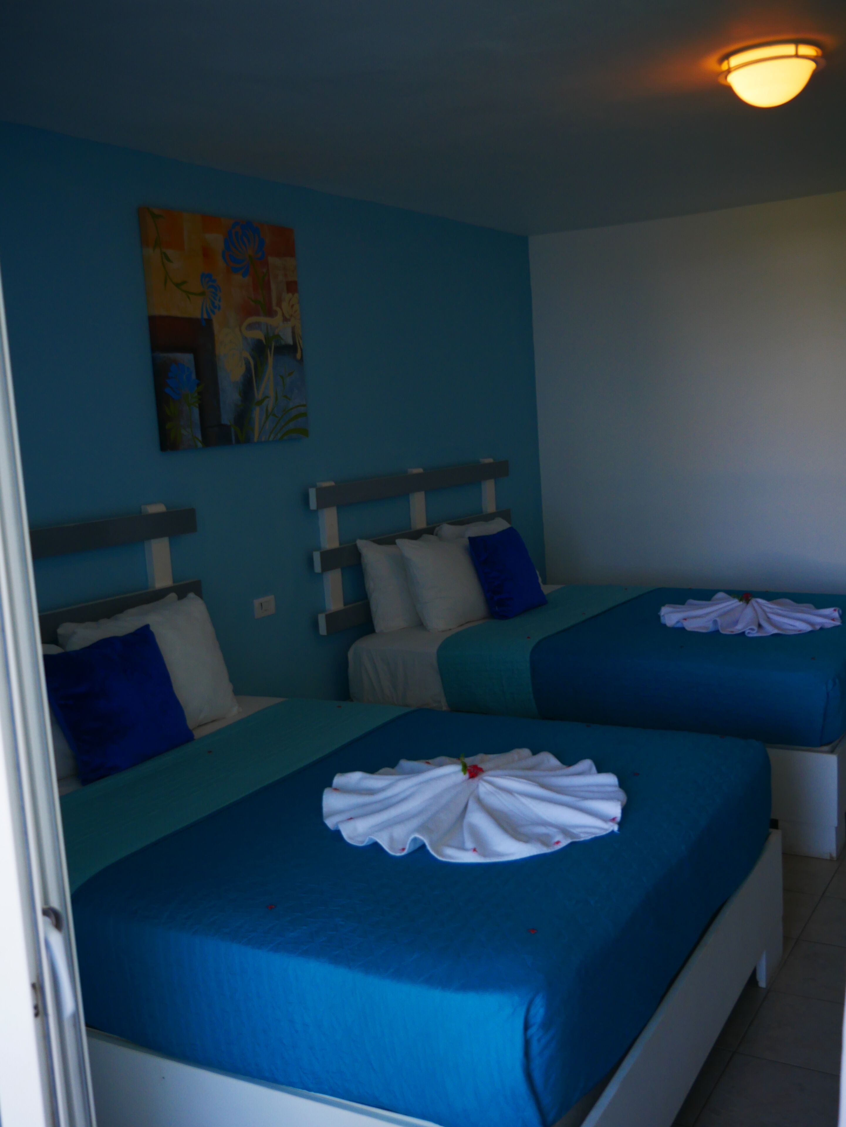 A hotel room with two beds, each with blue bedding and white pillows. There is a painting on the wall above the beds.