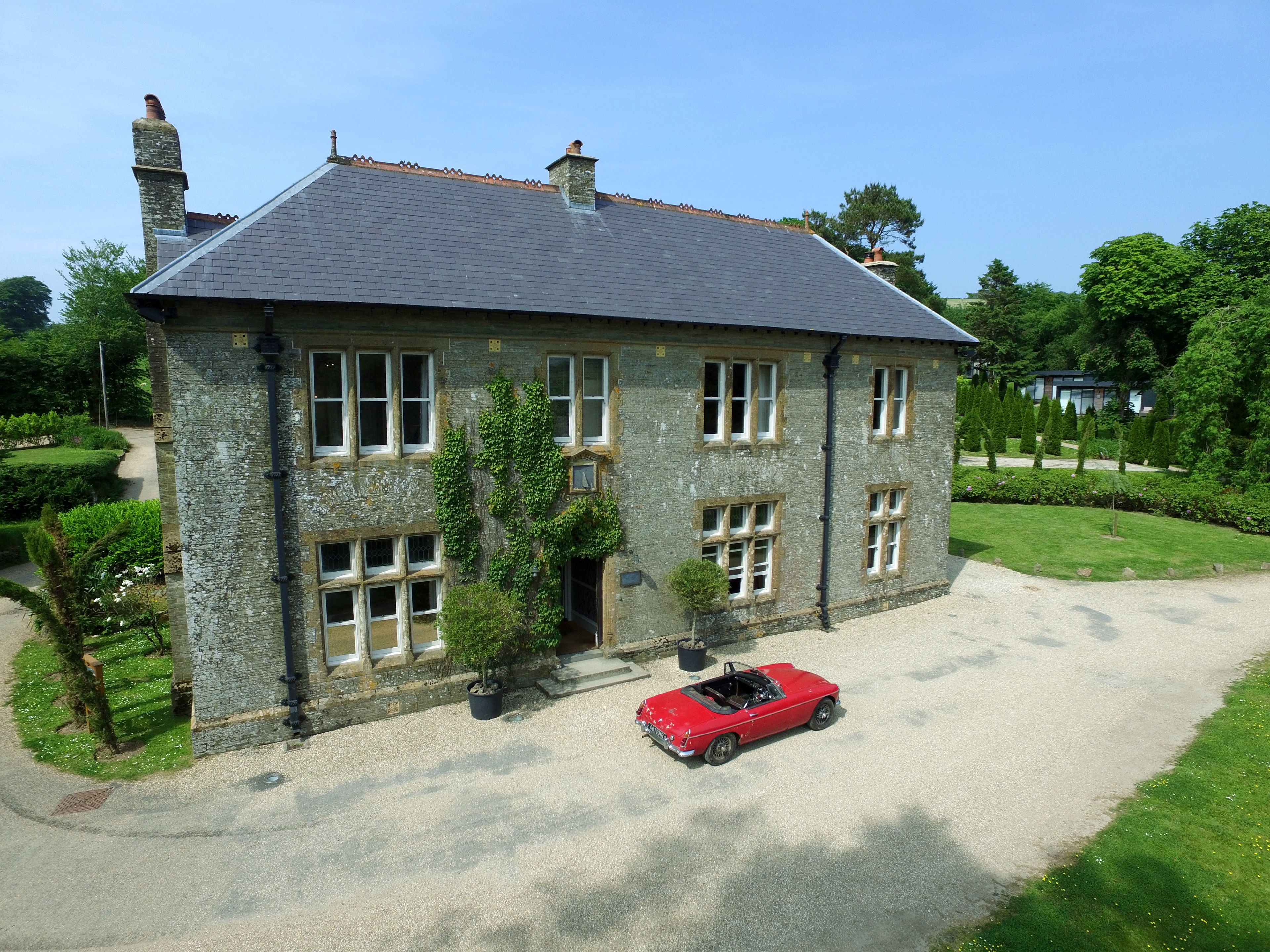 A stone building with a grey roof, a red convertible, and a well-maintained garden.