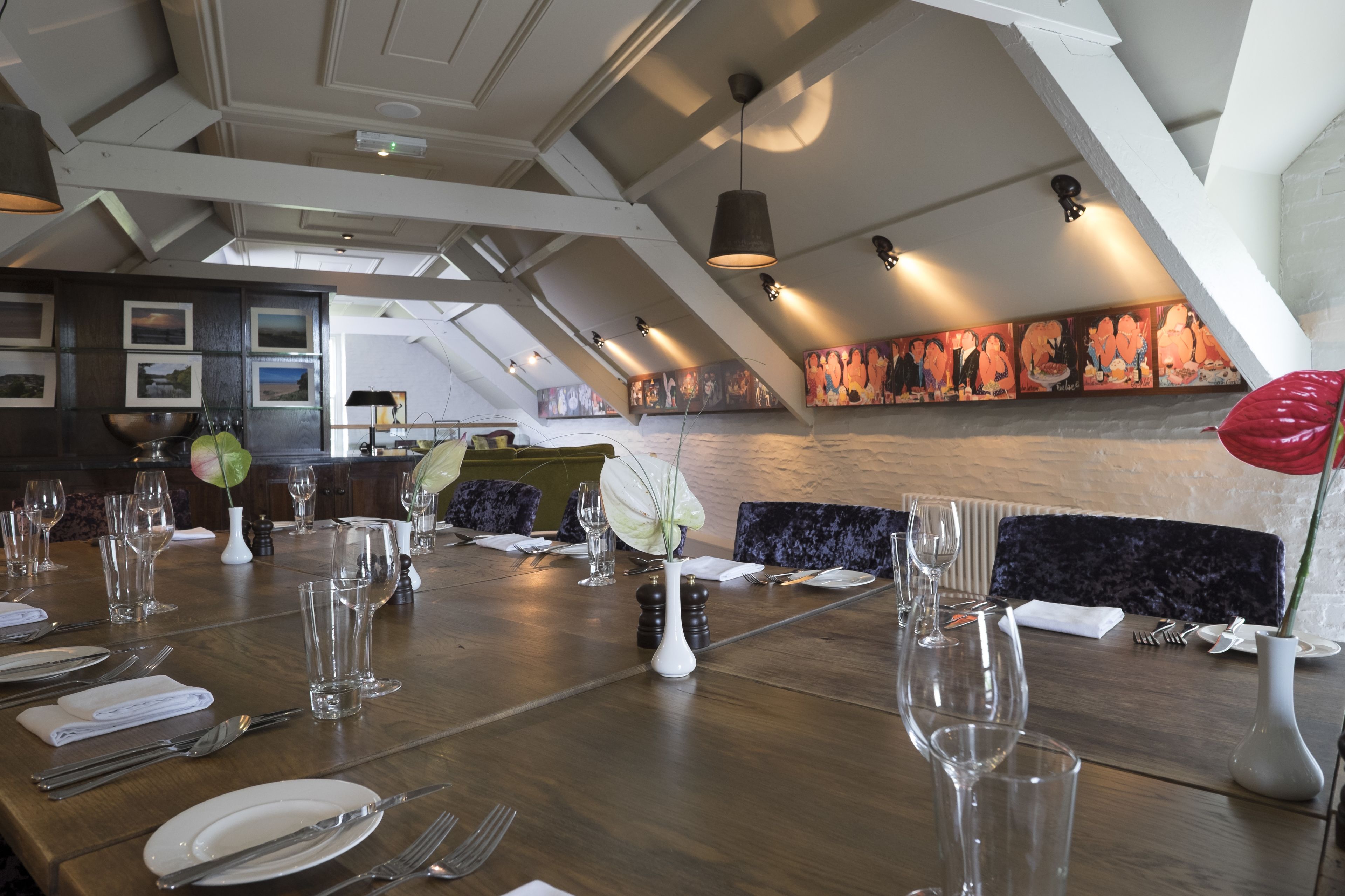 A dining area with a long wooden table set for a meal, featuring clear glassware and plates, a vase with a flower, and a wall with colorful artwork.
