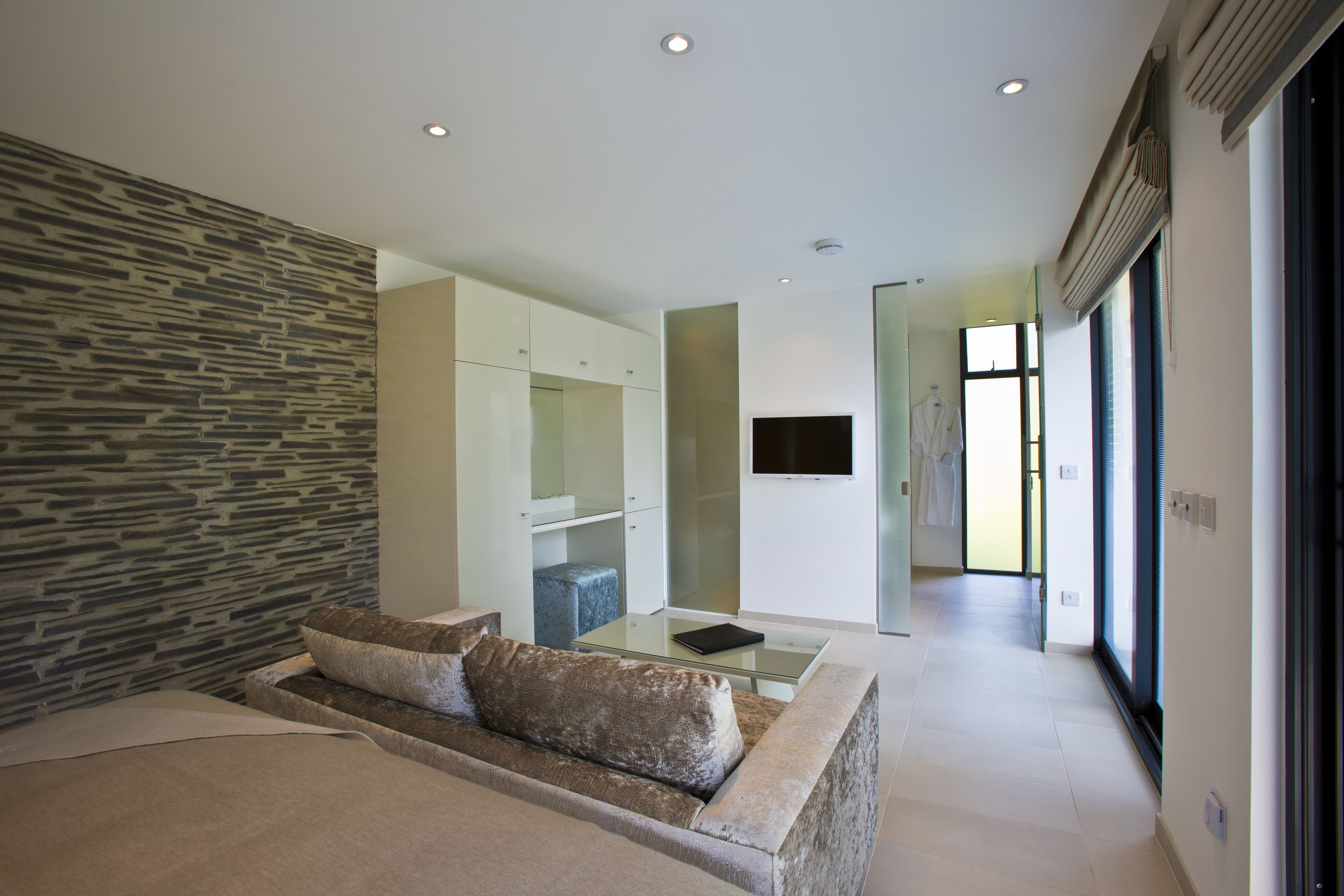 A modern hotel room with a stone accent wall, a glass coffee table, a sofa with cushions, a flat-screen TV, and a view of the hallway.