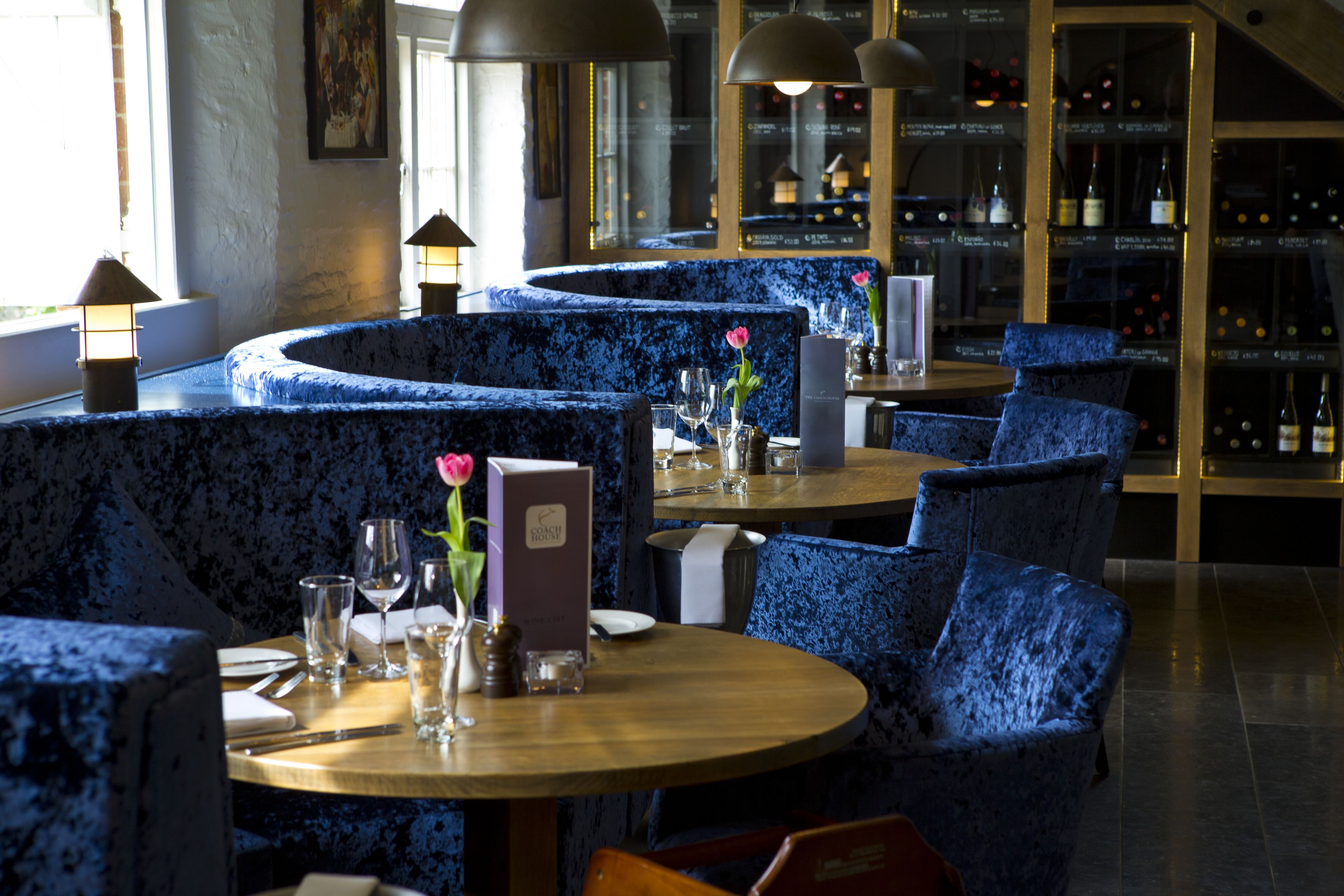 Een restaurant met blauwe bekleden zitjes, ronde tafels en een wijnrek op de achtergrond.