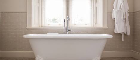 Une baignoire blanche, autoportante, dans une salle de bains aux murs recouverts de carreaux beiges et dotée d’une fenêtre avec des stores.