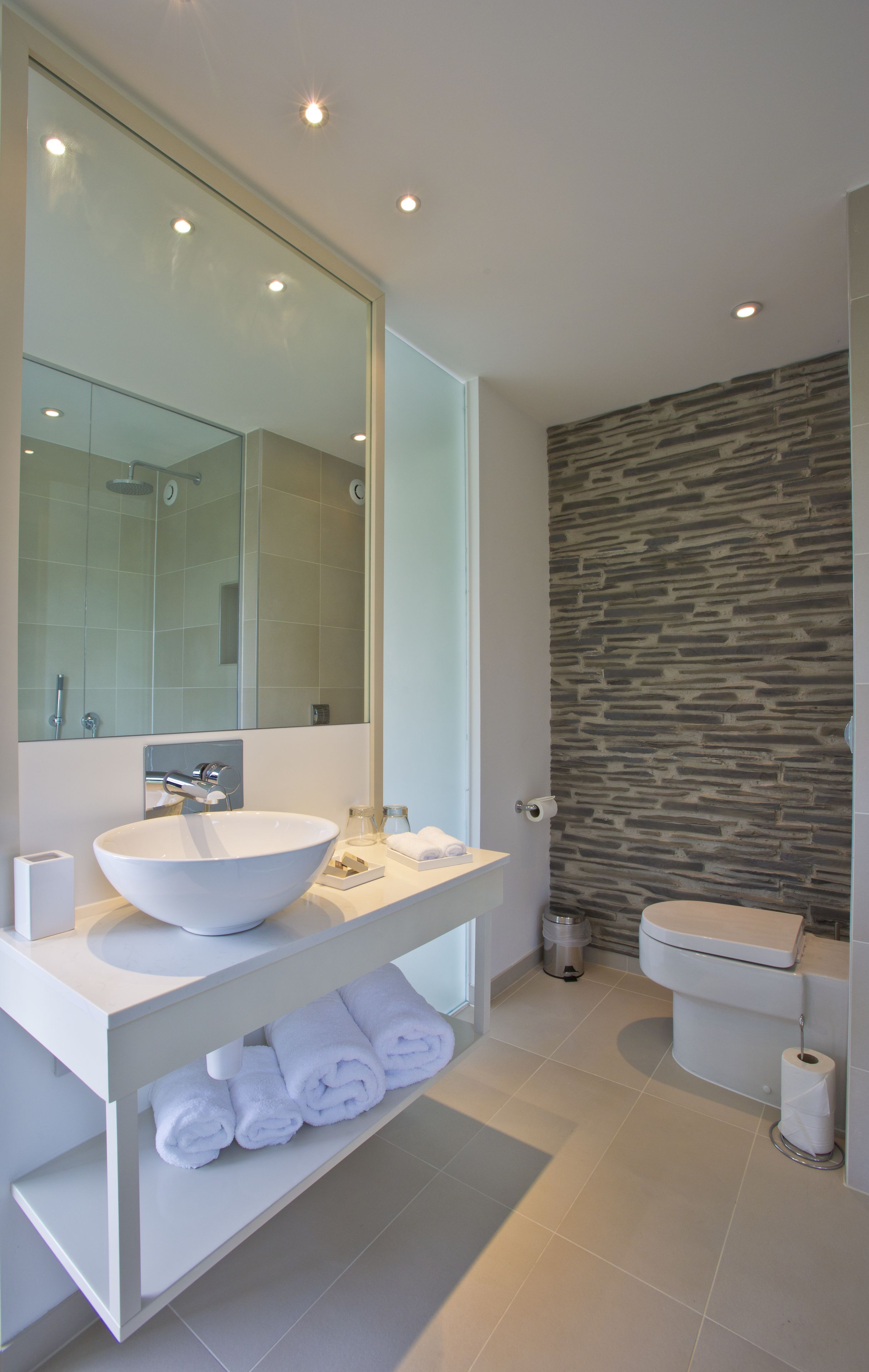 A modern bathroom with a large mirror, white sink, and a stone-textured wall.