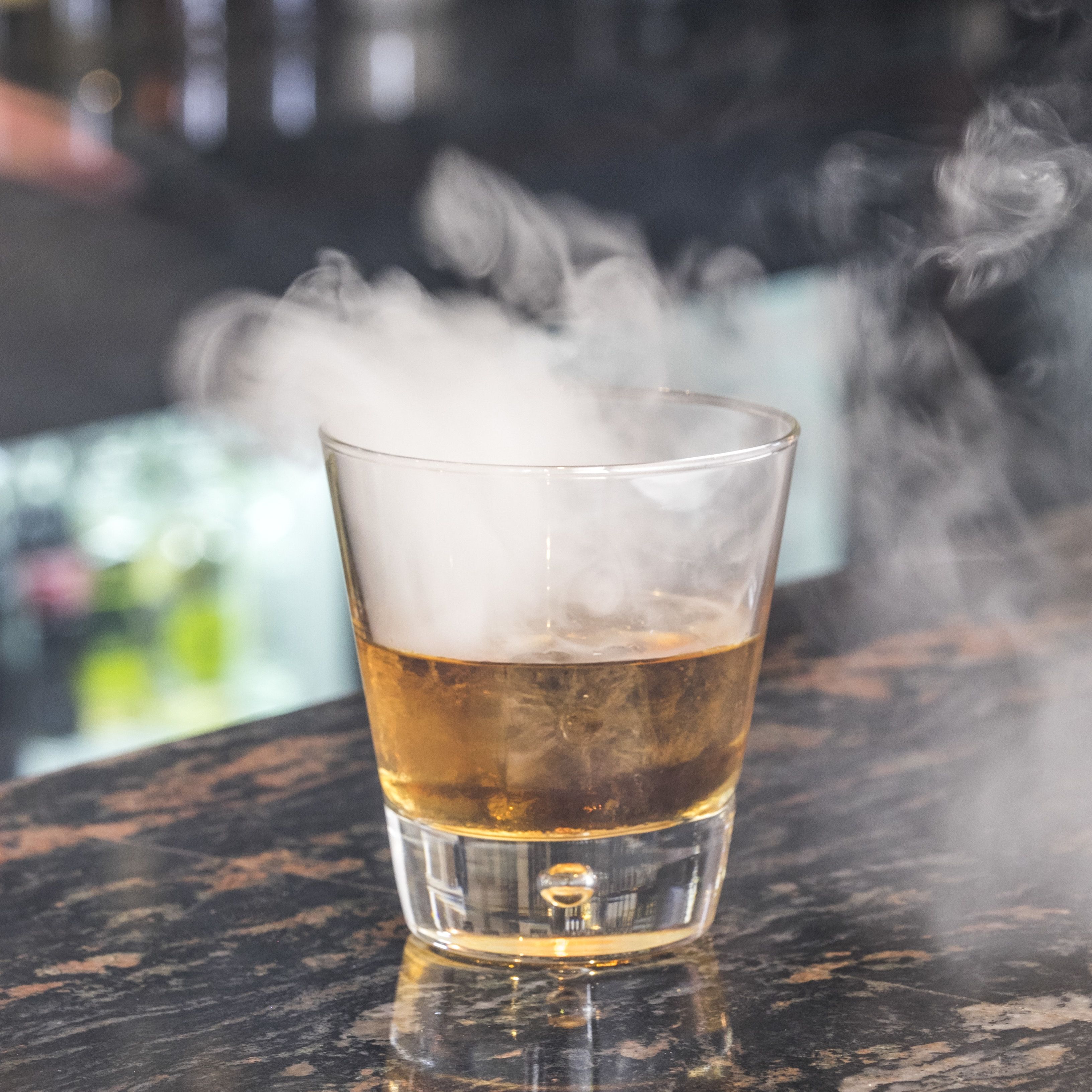 Un verre contenant un liquide ambré, surmonté d’une vapeur fumée, posé sur une surface sombre et texturée.
