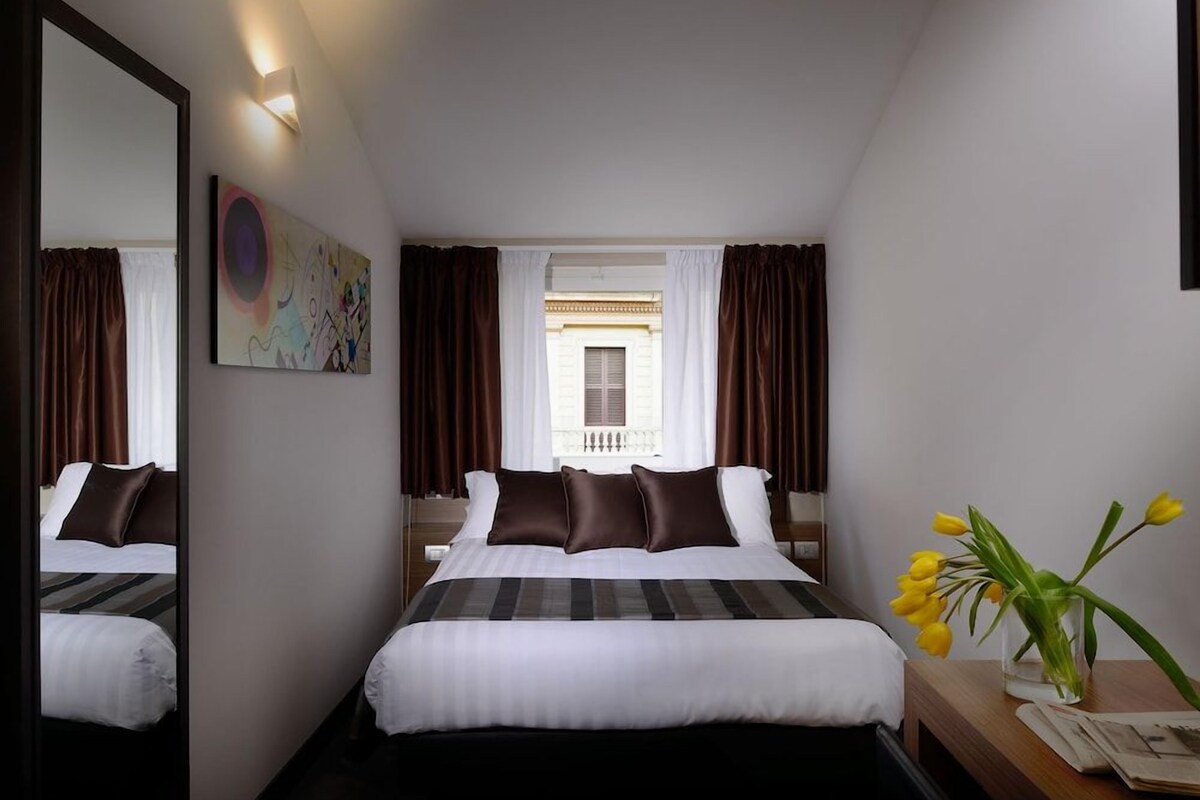 Habitación de hotel con una cama grande, una mesita de noche con un jarrón de flores, una ventana con cortinas y un cuadro en la pared.