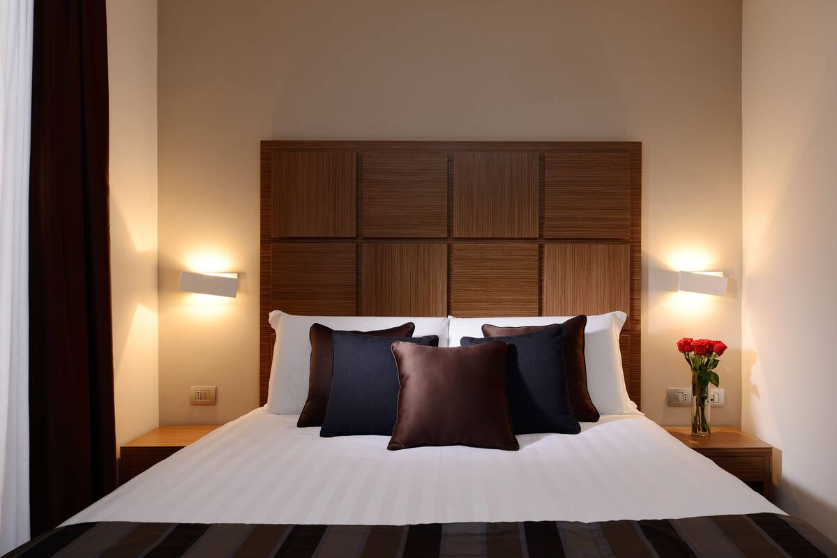 Une chambre d’hôtel avec un grand lit, une tête de lit en bois, deux tables de chevet et une petite table de nuit ornée d’un vase rempli de fleurs rouges.
