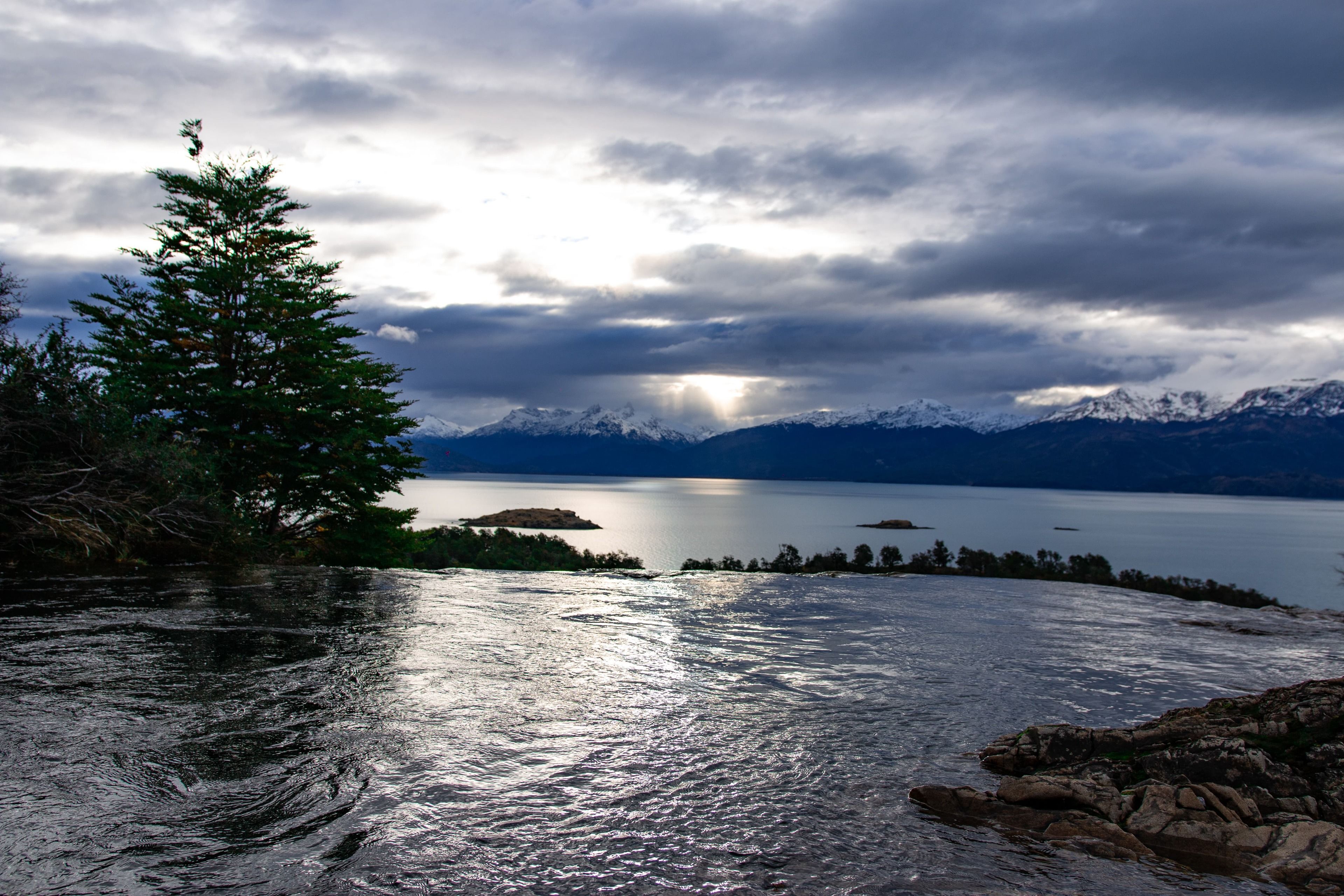 A calm lake with mountains in the background and a tree on the left.