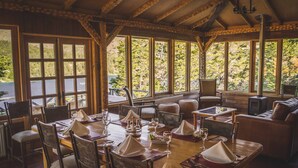 A cozy dining area with wooden furniture, large windows, and a stone fireplace. - Parador Austral Lodge (Chile Chico)