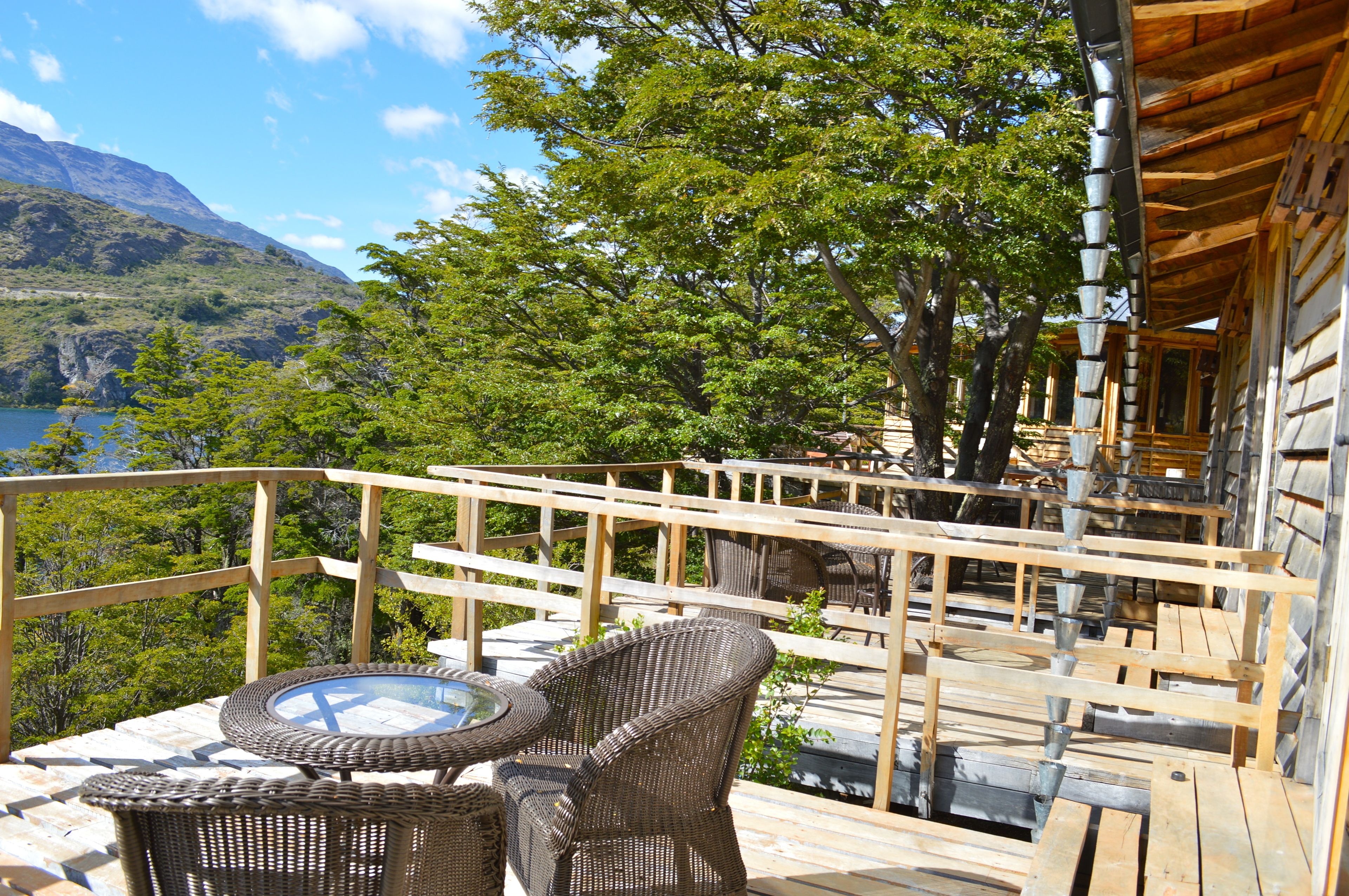 A wooden deck with wicker furniture and a glass table, overlooking a mountainous landscape and a body of water.