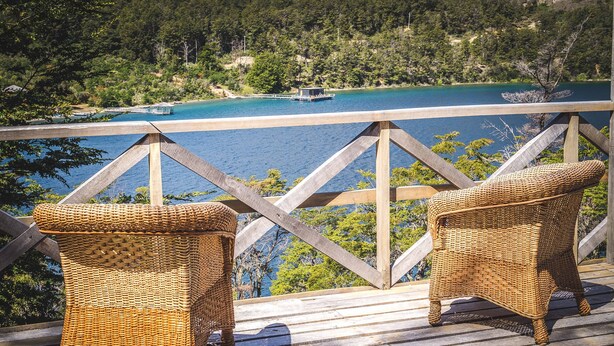 A wooden deck with two wicker chairs overlooking a lake with a boat and dock. - Parador Austral Lodge (Chile Chico)