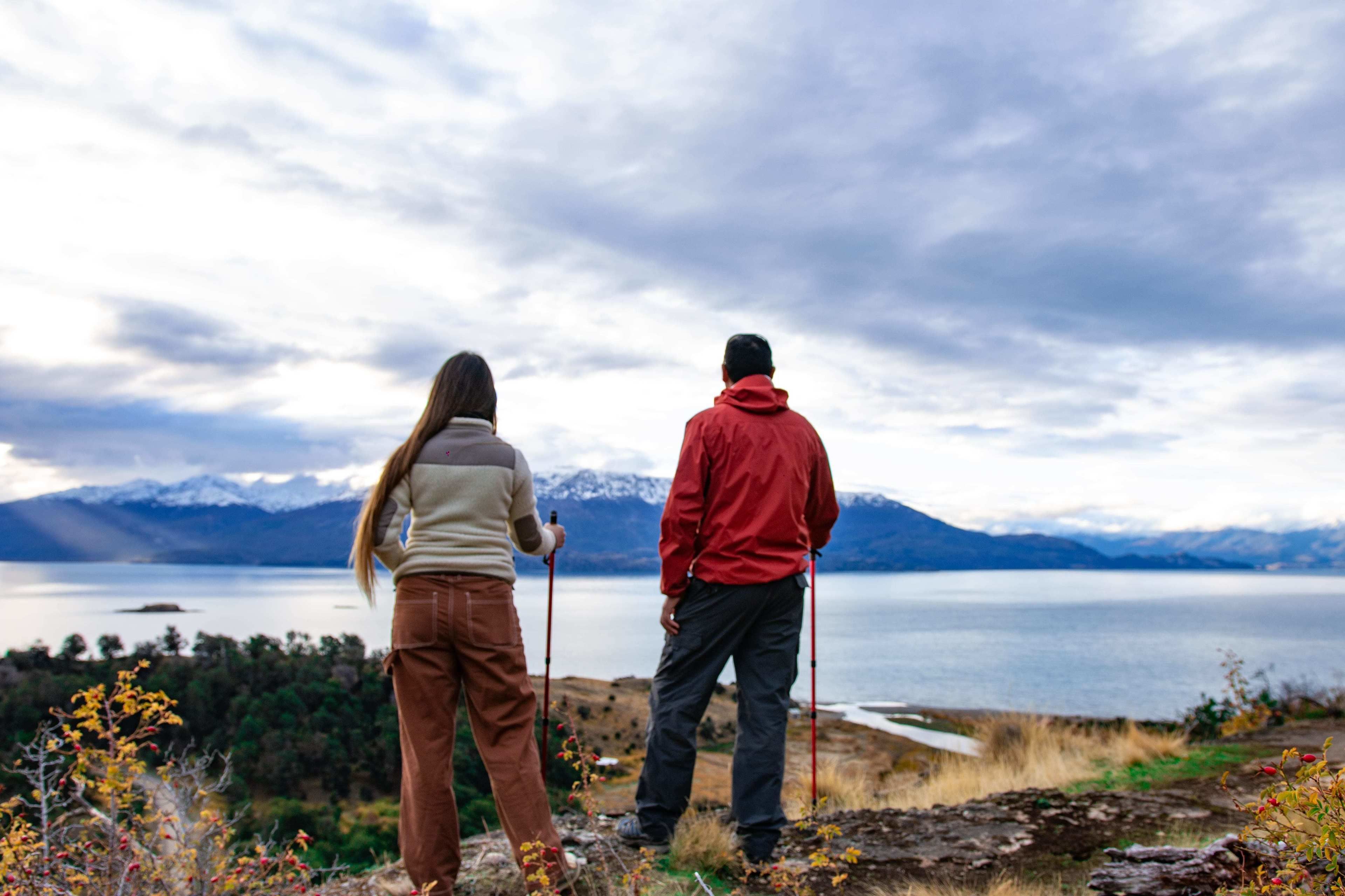 Two people standing on a rocky shore with a lake and mountains in the background.