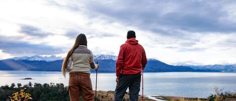 Two people standing on a rocky shore with a lake and mountains in the background.