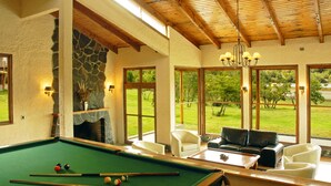 A pool table with balls, a fireplace, and a view of a green landscape through large windows. - Parador Austral Lodge (Chile Chico)