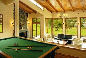 A pool table with balls, a fireplace, and a view of a green landscape through large windows.